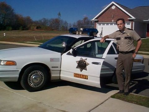 1997 Ford Crown Victoria - A sheriff stands by a white 1997 Ford Crown Victoria police car parked in a driveway.