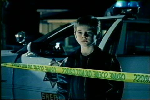 1997 Ford Crown Victoria - A young boy stands in front of a police car, separated by crime scene tape.