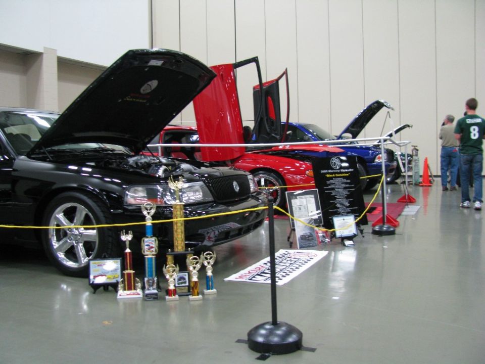 Black Mercury Marauder with hood open and trophies in front, next to a red car with gullwing doors