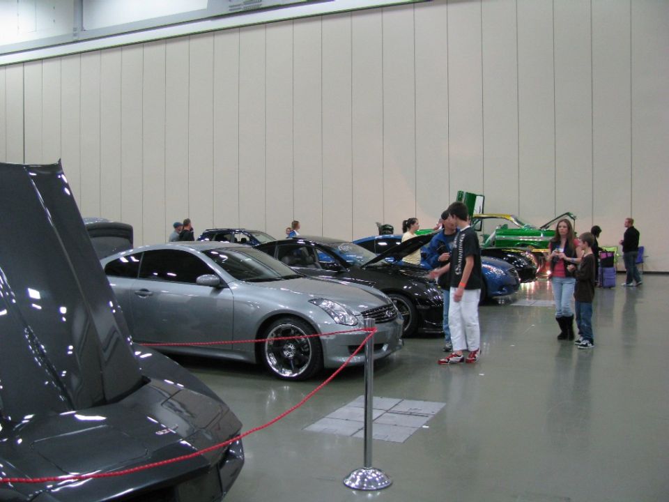 Several cars are on display indoors, with people looking at them. A gray coupe is in the foreground
