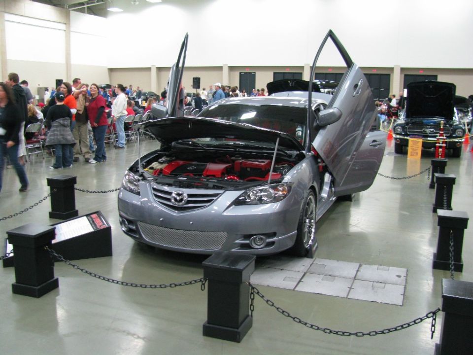 Gray Mazda 6 with hood and gullwing doors open, showing red engine components. Another car is visible in the background