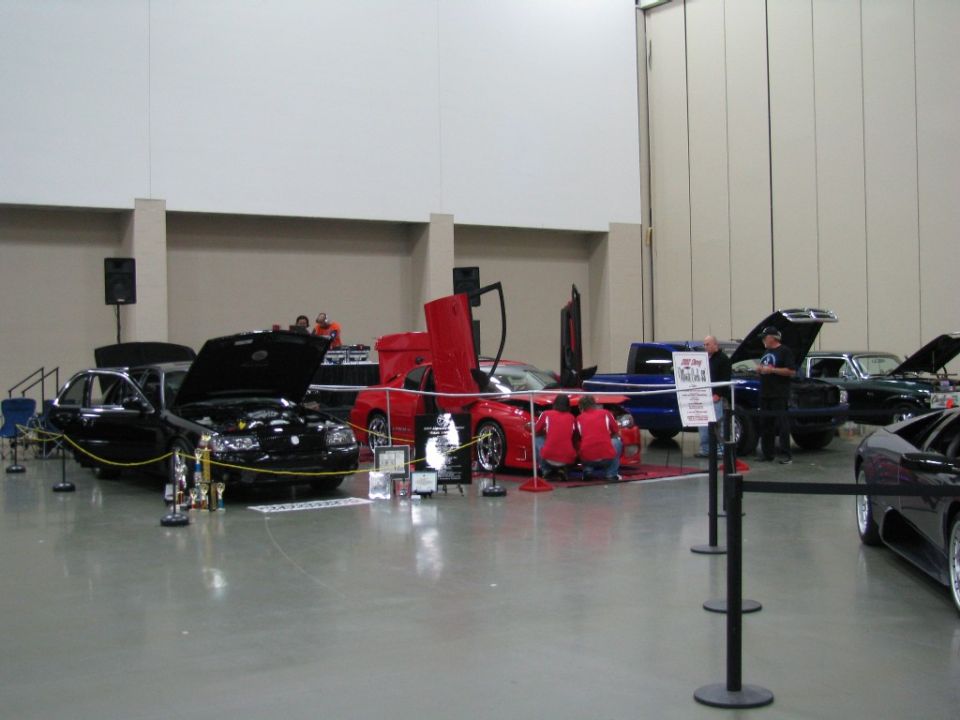 Black sedan with hood open, red sports car with scissor doors, and blue pickup truck at an indoor car show