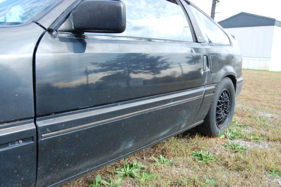 Side view of a dark-colored car's door and rear wheel with a mesh-style rim