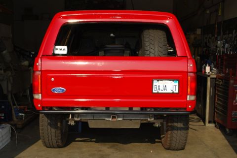 1986 Ford Bronco - Rear view of a red Ford Bronco SUV inside a garage, showing the tailgate with a Ford oval emblem, a PIAA sticker, and a California license…