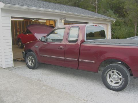 2007 Ford Mustang - A maroon pickup truck is parked in front of a garage, with another vehicle visible inside with its hood open.