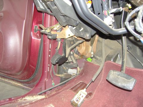2007 Ford Mustang - Driver's footwell of a burgundy-interior vehicle with the dash removed, showing the parking brake pedal assembly with 'BRAKE' release handl…