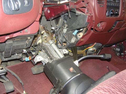 2007 Ford Mustang - Burgundy truck interior mid-teardown, with the lower dash panels off and the steering column dropped onto the seat still wearing its black…