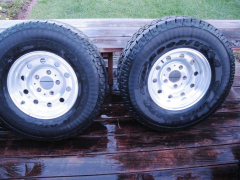 1986 Ford Bronco - Two Goodyear Wrangler tires mounted on polished aluminum wheels are displayed on a wet wooden surface.
