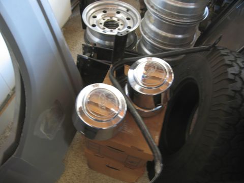 1986 Ford Bronco - Two chrome Ford hubcaps sit on top of stacked wheels and tires in a garage.