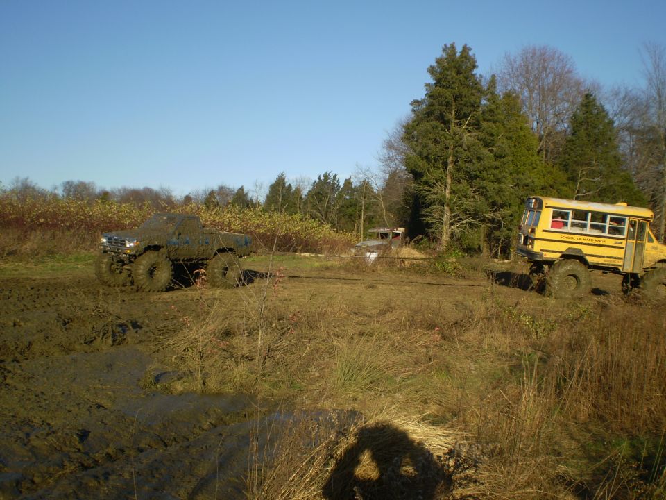 Lifted muddy pickup and yellow 'School of Hard Knox' off-road short bus in a rutted mud field