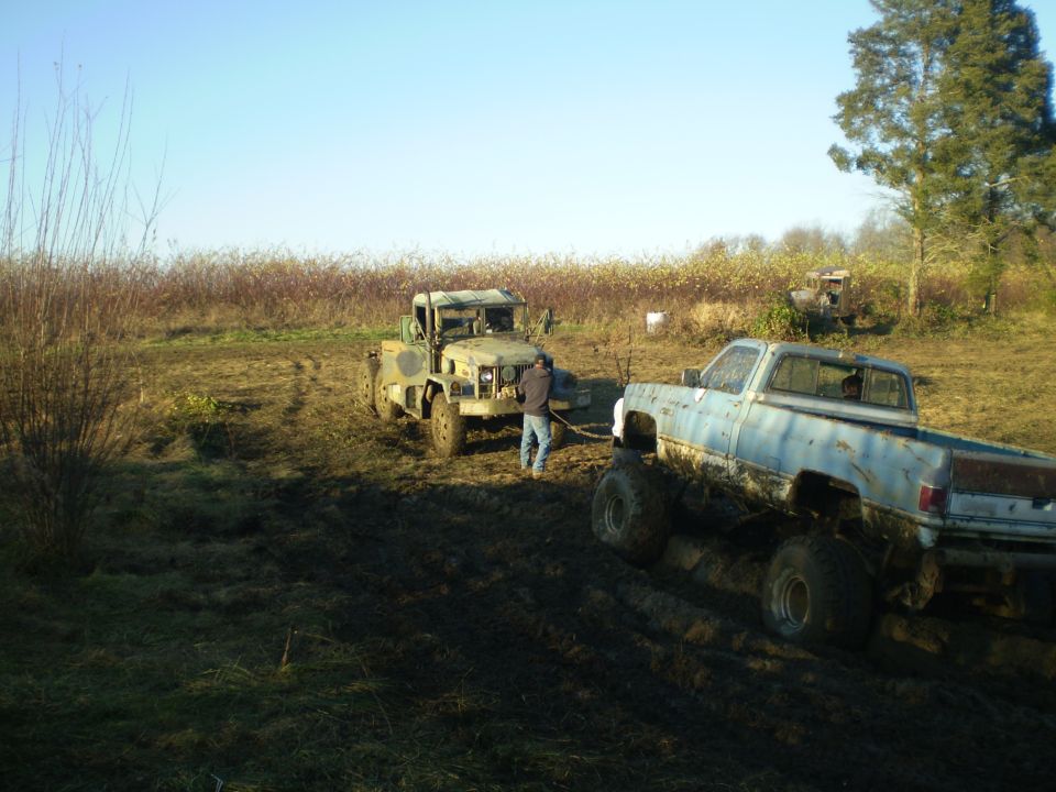 Muddy field with a light blue pickup truck and a military-style truck stuck in the mud, a person is between them