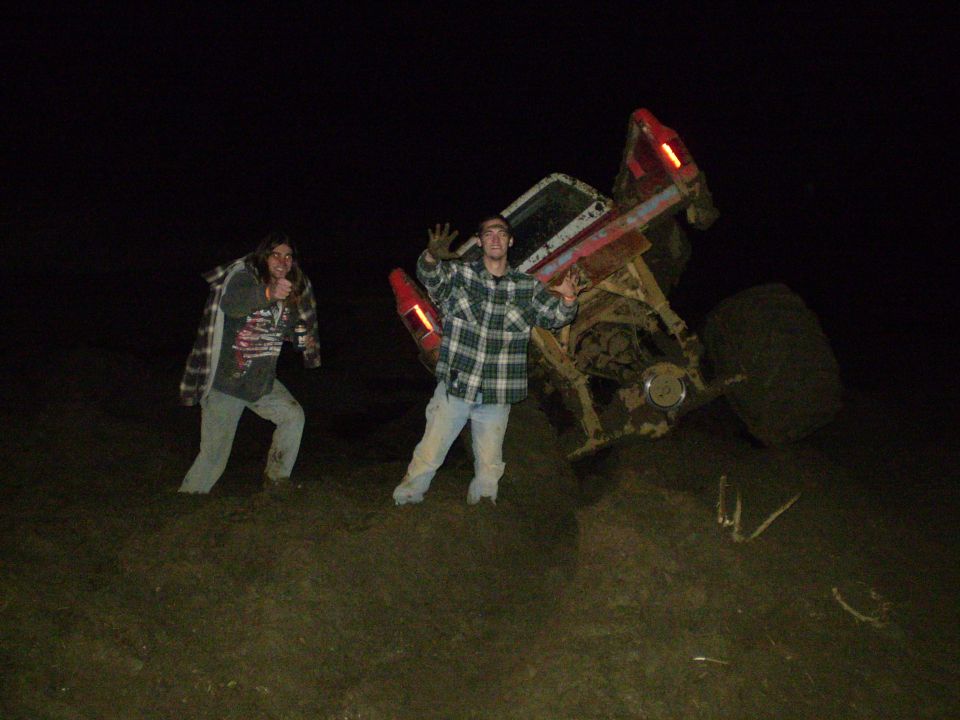 Overturned red truck in mud at night with two men standing nearby