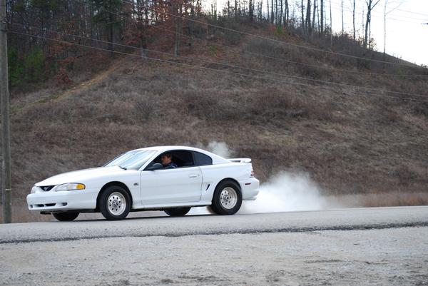white coupe doing a burnout on a road with trees in the background