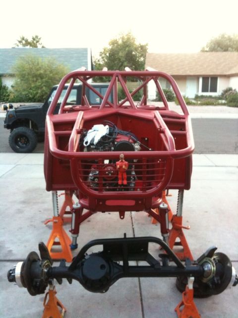 Suzuki Samurai - Front view of a red tube-frame vehicle chassis with roll cage, mounted on orange jack stands in a driveway, with a black solid rear axle as…