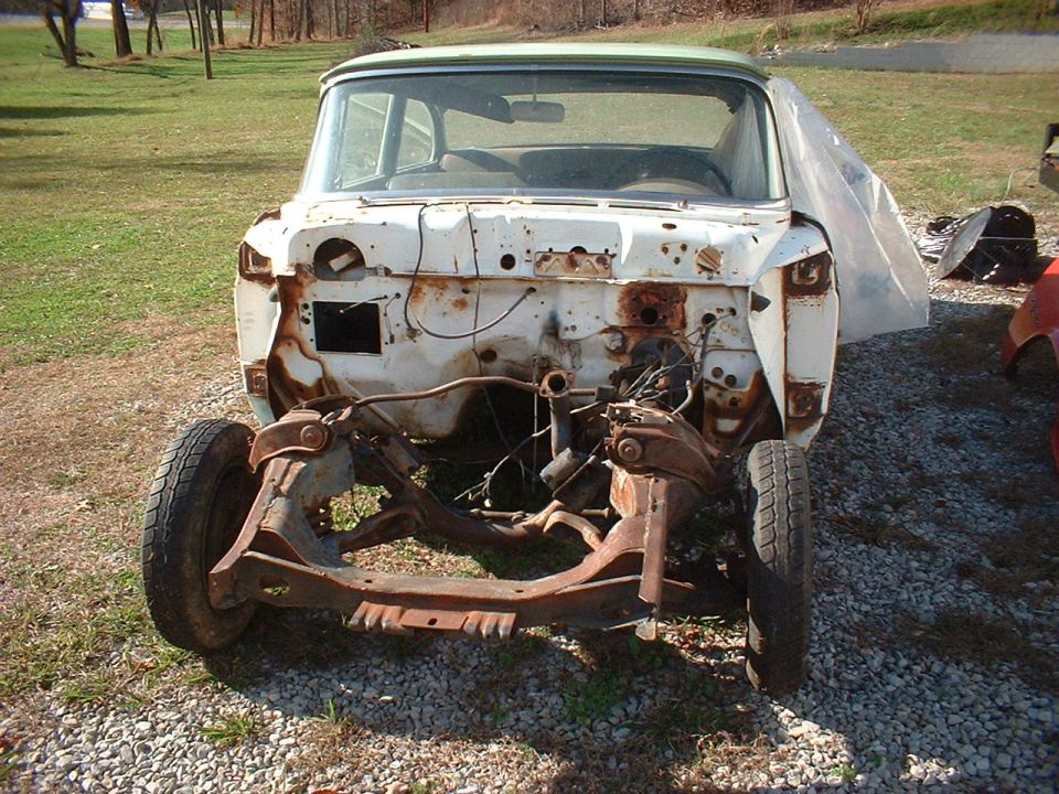 Stripped white sedan body shell with rusty firewall and exposed front frame on gravel