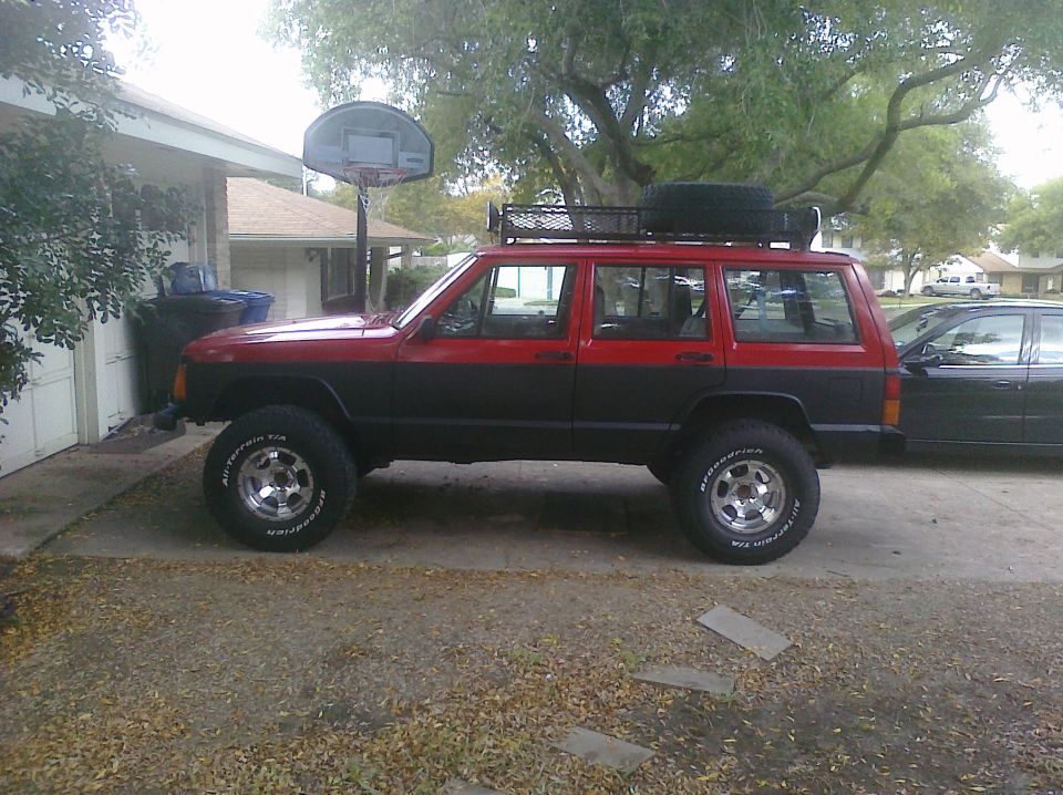 red and black Jeep Cherokee with roof rack and spare tire on top, parked next to a black sedan