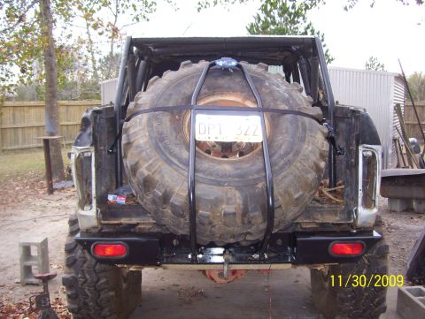 1978 Ford Bronco - Rear view of a stripped topless 4x4 with a fabricated black tubular swing-out tire carrier holding an oversized muddy bias-ply spare on a s…