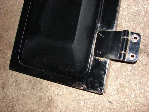 1984 Ford F-250 - A black vehicle door part with a metal hinge is shown against a concrete surface.