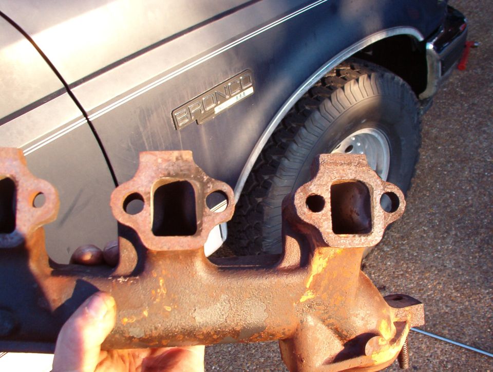 Rusty cast-iron exhaust manifold held in front of a gray Ford Bronco