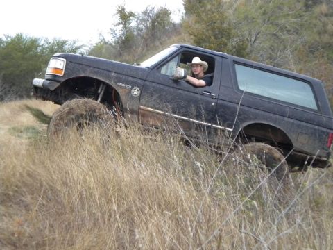 1995 Ford Bronco - A driver in a cowboy hat navigates a muddy, grassy incline in a black Ford Bronco.