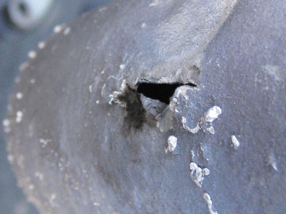 close-up of a damaged metal part with a hole and peeling paint, supermotors.net logo visible
