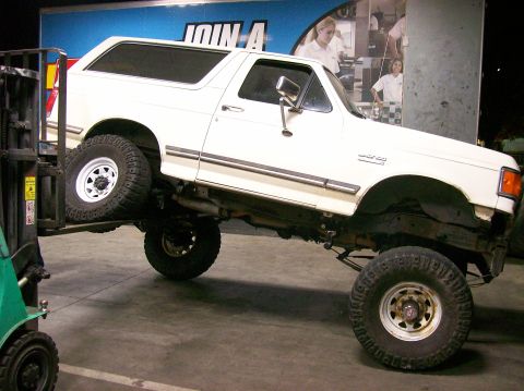 1989 Ford Bronco - A white Ford Bronco is being lifted by a forklift, showing its undercarriage and lifted suspension.