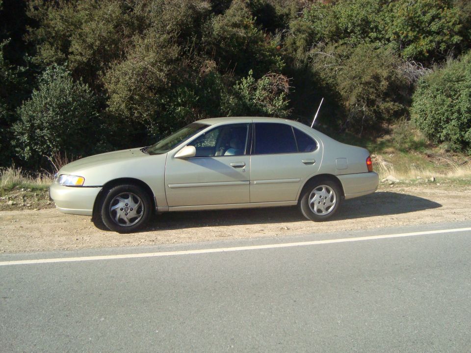 tan sedan parked on dirt shoulder next to a road, with trees and a hill in the background
