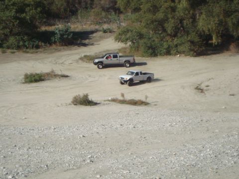 1986 Ford Bronco - Two pickup trucks navigate a dusty, off-road path surrounded by trees.