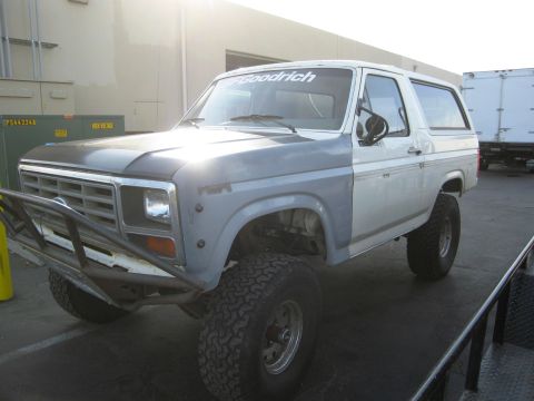 1986 Ford Bronco - A silver and white two-door Ford Bronco SUV with a matte black hood, lifted on BFGoodrich mud-terrain tires with chrome wheels, parked outd…