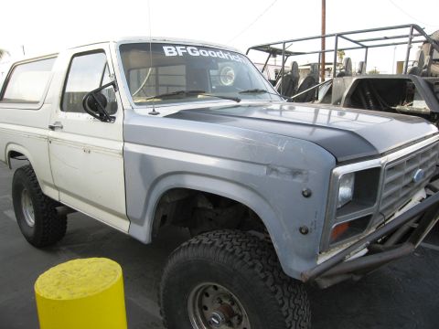 1986 Ford Bronco - A two-tone white and gray 1980s Ford Bronco SUV shown from a 3/4 front angle, with a black hood, oversized BFGoodrich all-terrain tires on…