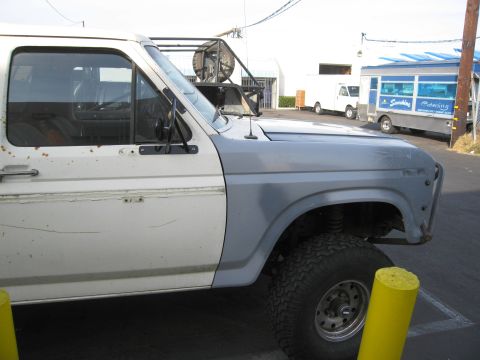1986 Ford Bronco - This Ford Bronco has a two-tone paint scheme with a gray front fender and white body. A catering truck and a white van are in the backgroun…