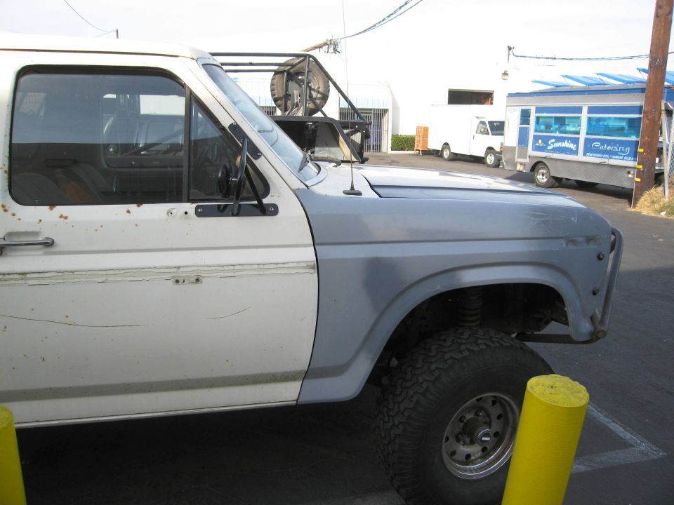 Side view of a white Ford Bronco with a gray fender and off-road tires. Two other vehicles are visible in the background