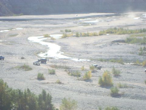 1986 Ford Bronco - Multiple trucks are seen driving through a dry riverbed with a small stream meandering through the rocky terrain.