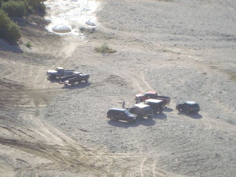 1986 Ford Bronco - Multiple trucks and SUVs gathered in an off-road area with dry creek bed in the background.