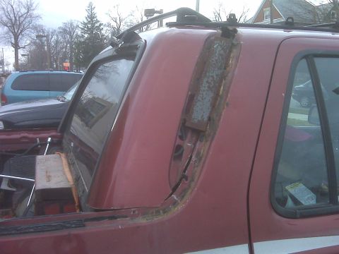 1991 Ford Bronco - The rear passenger side of a red SUV shows significant damage to its roof trim and window seal, with exposed metal and rust.