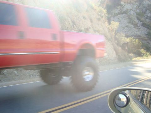 1986 Ford Bronco - Red pickup truck with large tires driving on a road next to a hill.