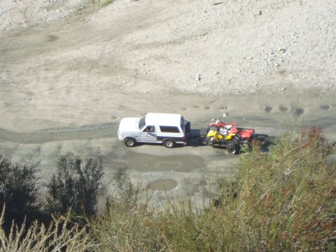 1986 Ford Bronco - A white SUV tows a trailer loaded with ATVs along a dirt path, with brush in the foreground.