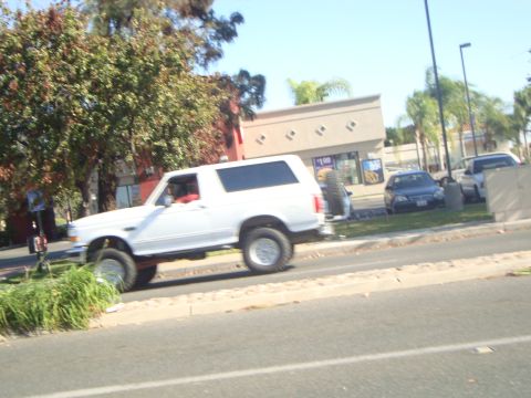 1986 Ford Bronco - A white SUV with a lifted suspension and large tires drives down a street.