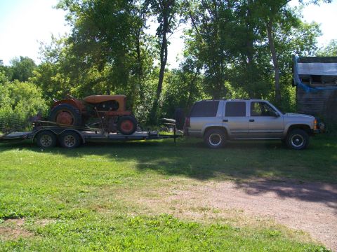2000 Chevrolet Tahoe - Silver SUV towing a trailer with an orange tractor on a grassy area with trees in the background.