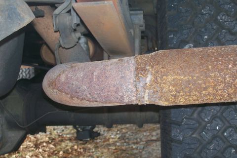 1992 Ford C-350 (Centurion Conversions) - The underside of a vehicle shows a heavily rusted exhaust pipe connected to what appears to be a muffler. Leaf springs and a tire are also…