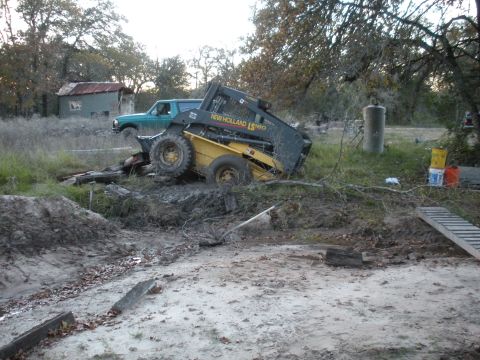 1996 Ford Bronco - A green pickup truck and a New Holland skid steer loader are stuck in muddy terrain near a rural building.