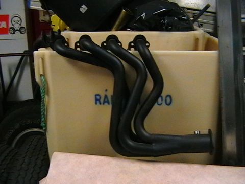 1996 Ford Bronco - A black exhaust manifold with four pipes is shown, resting against a tan container with blue lettering.