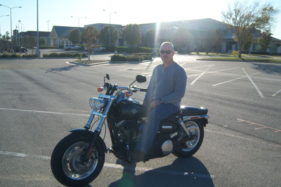 Man in sunglasses and a blue shirt sits on a black motorcycle in a parking lot. Buildings and trees are in the background