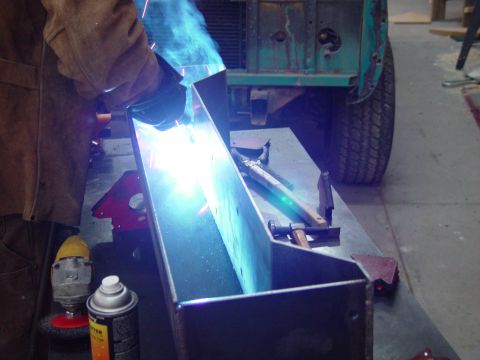 1993 Ford Bronco - A person welds a metal component in a workshop, with the fender and tire of an older green vehicle visible in the background.