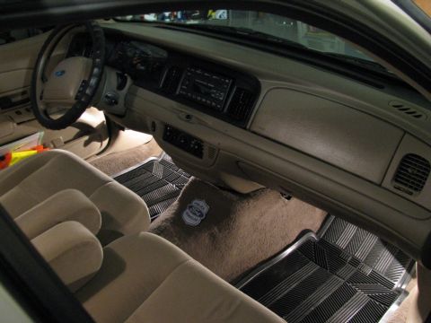2004 Ford Crown Victoria - The interior of a 2004 Ford Crown Victoria, tan upholstery and a police-themed floor mat, is visible from the passenger side.