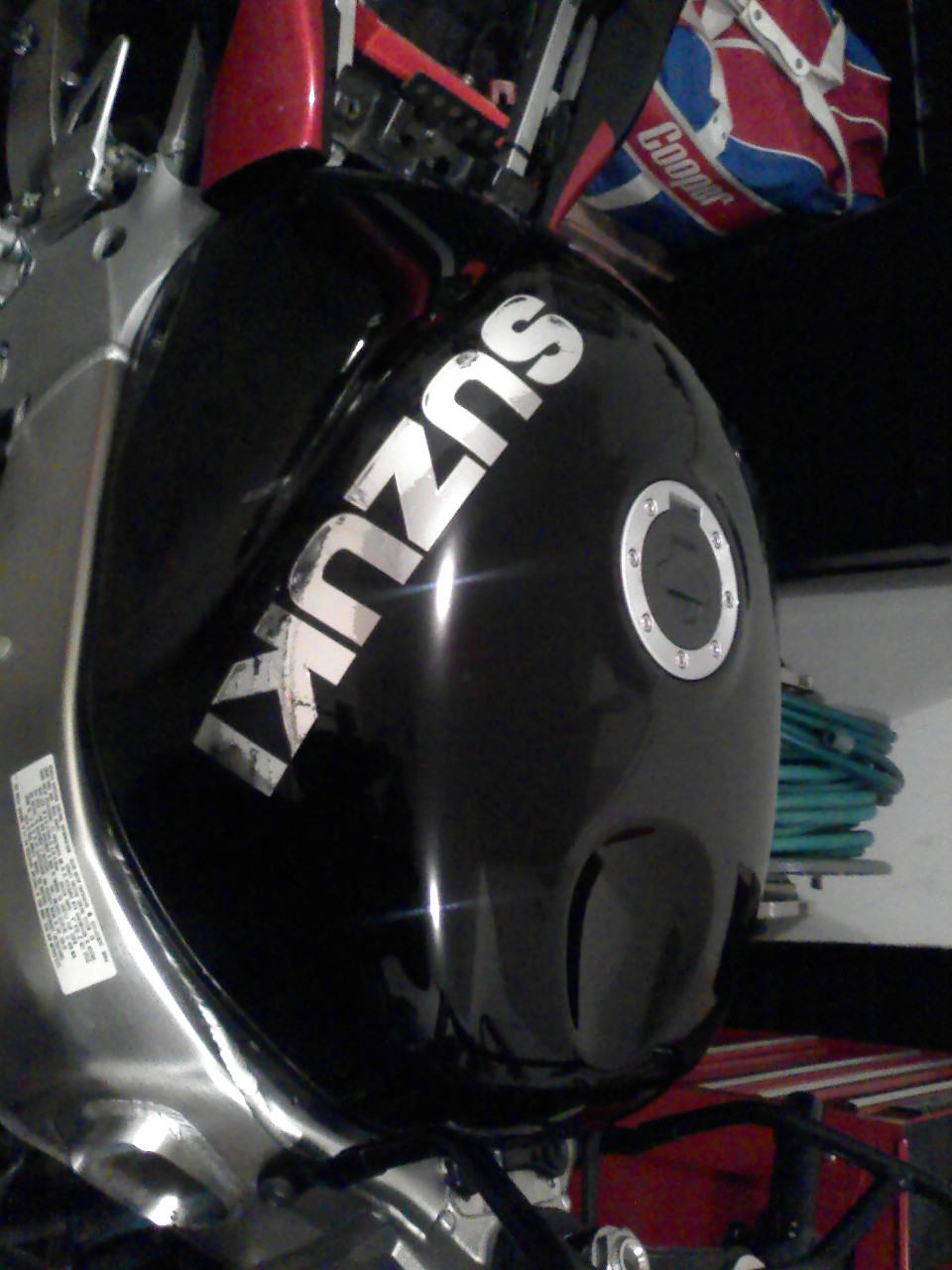 Black Suzuki motorcycle fuel tank with worn silver SUZUKI decal and fuel cap, photographed sideways in a garage