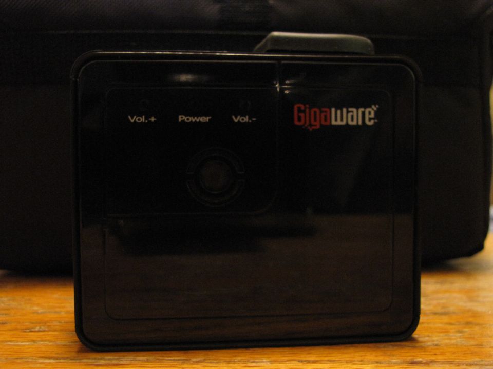 Black GigaWare projector with 'Vol.+', 'Power', and 'Vol.-' buttons on a wooden surface