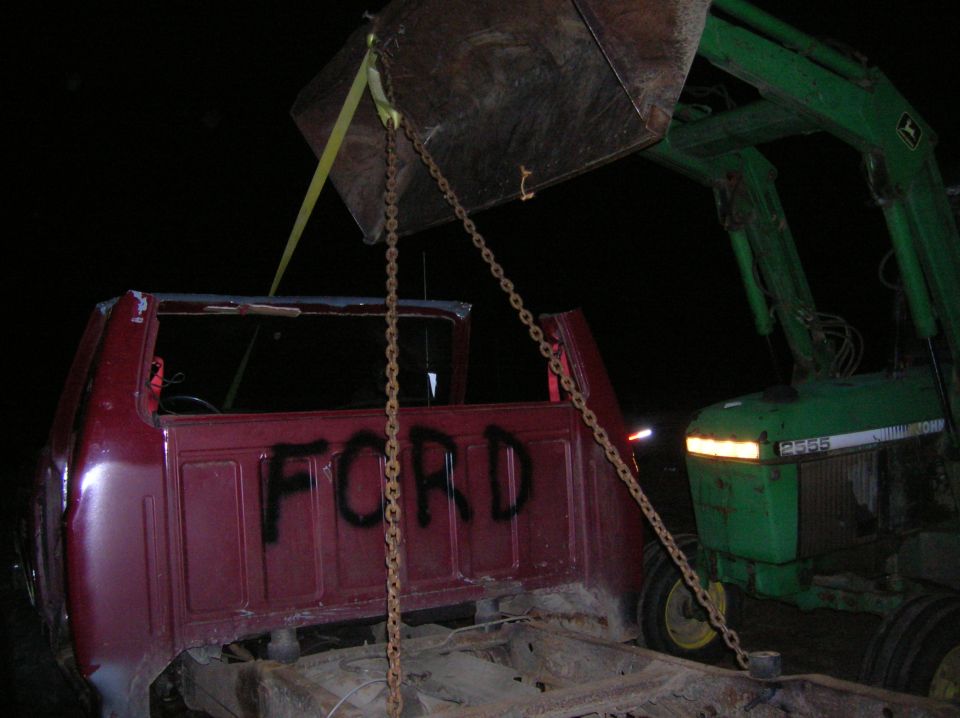 Ford truck cab and chassis being lifted by a green tractor with a front loader