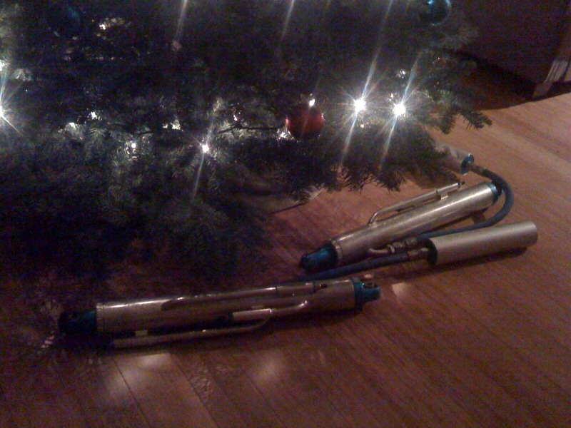 Two silver fuel pump assemblies with blue connectors and black hoses on a wooden floor near a Christmas tree