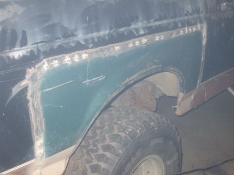 1995 Ford Bronco - Rust repair in progress on a truck's rear quarter panel: a large teal-green patch panel has been welded in with a continuous row of plug we…
